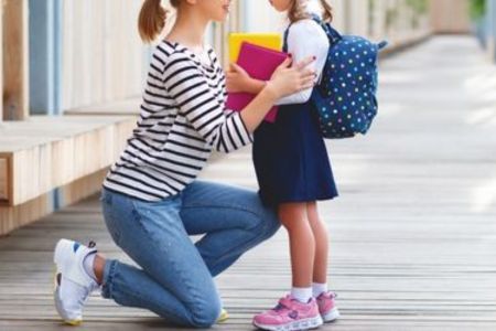 Vrouw die op haar knieen zit en een meisje vasthoudt dat een rugzak op heeft en schoolspullen in haar handen heeft