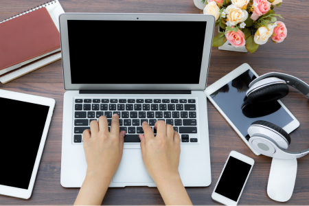 Twee handen die aan het typen zijn op een laptop, met allerlei digitale middelen eromheen